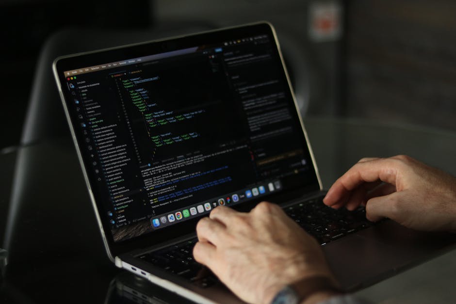 A close-up of a person's hand operating a silver laptop on a wooden surface, with the screen displaying lines of computer code in a text editor, featuring various syntax elements and highlighted text.