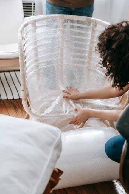 A person is wrapping a piece of clear plastic protective covering around a white woven material laundry basket in a home interior setting. The basket is positioned on a wooden floor near a white sofa,