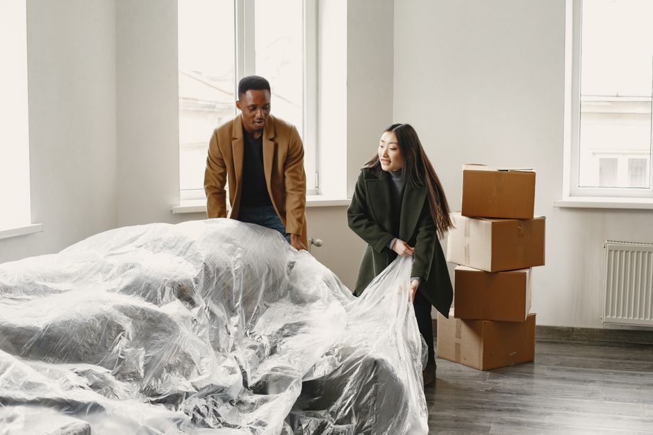 A person is wrapping a piece of clear plastic protective covering around a white woven material laundry basket in a home interior setting. The basket is positioned on a wooden floor near a white sofa,